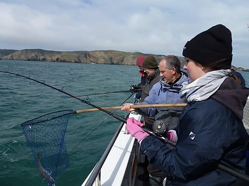 Two anglers at the rail netting a fish under steep green cliffs